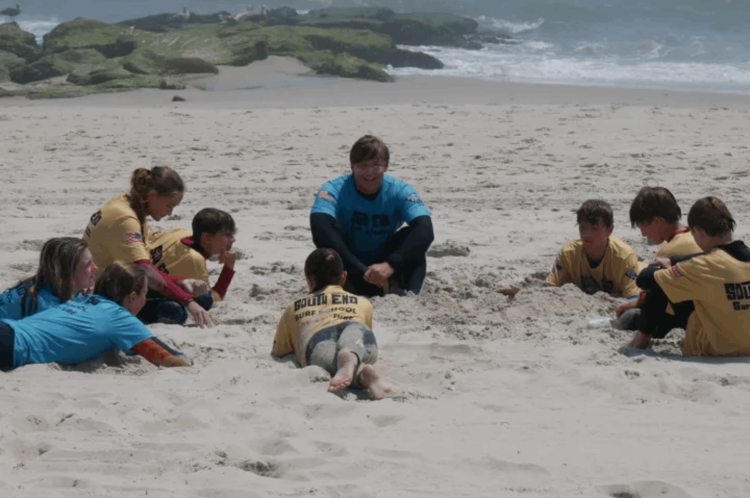 A group of children sit together in a circle on the beach, listening to their group surf instructor in Beach Haven, NJ.