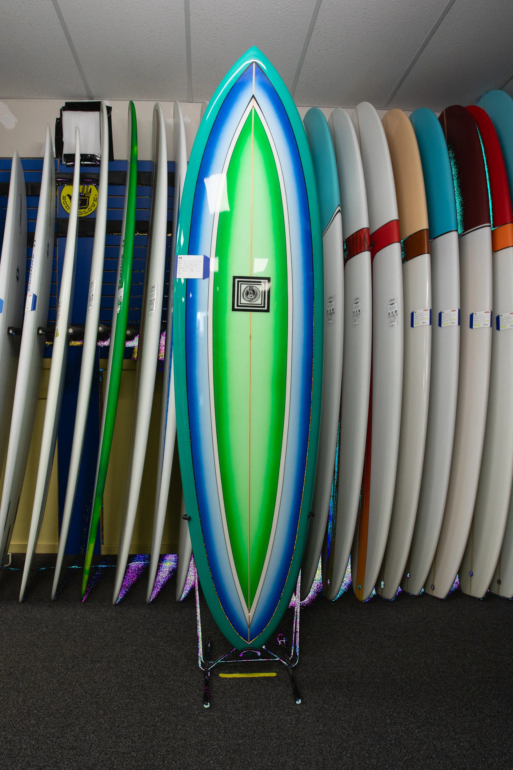 7’6” Clean Ocean Surfboard on the floor of LBI Surf Shop, South End Surf ‘N Paddle. The board is finished in a bright green and blue motif