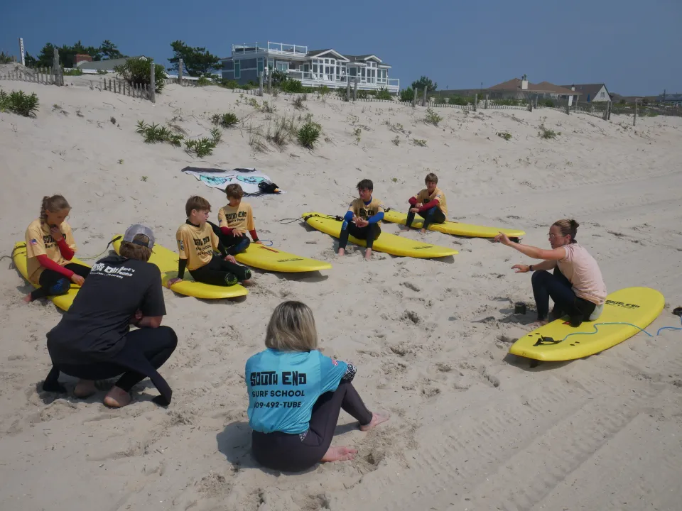 South End Surf School instructors demonstrating surfing techniques to a group of children with yellow surfboards on the beach