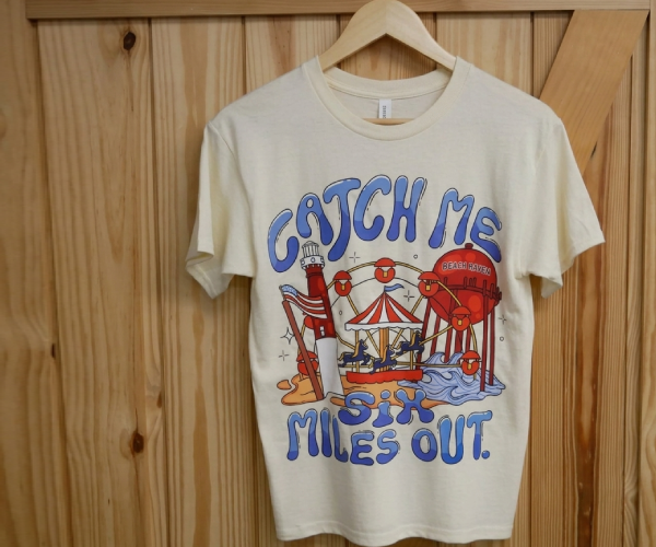 Custom Beach Haven shirt designed and sold by South End Surf ‘N Paddle displayed on a hanger. Design features a cartoon version of a pier and the Beach Haven water tower. Text displays “Catch Me Six Miles Out”