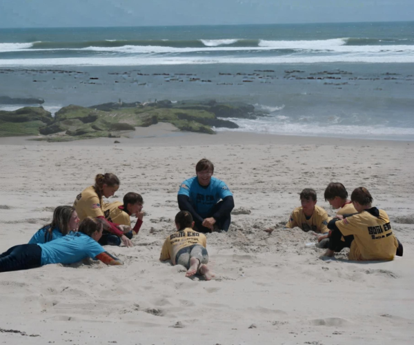 A group of children sit together in a circle on the beach, listening to their group surf instructor in Beach Haven, NJ.